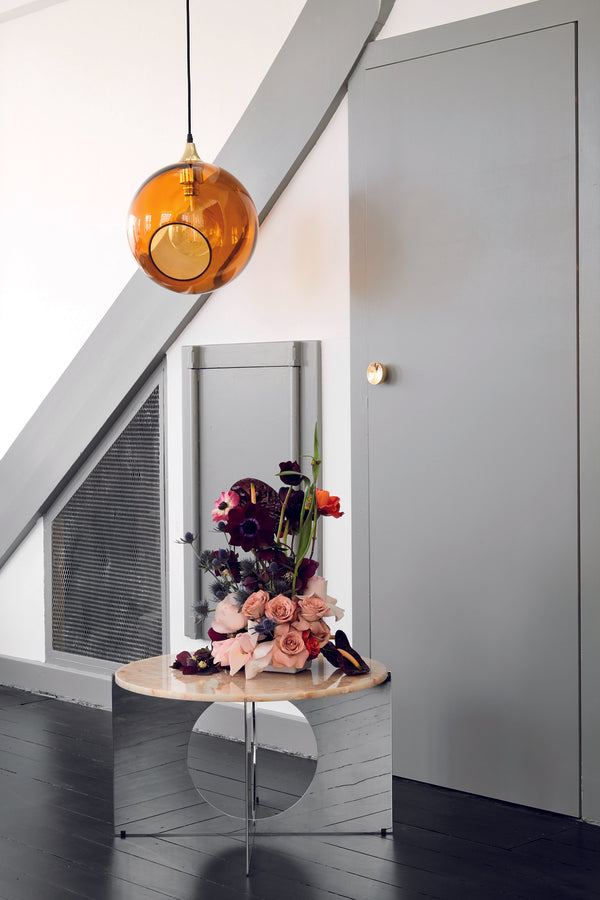 Rund lampa i orange/gult glas och med en avskuren bit med guldkant som ger en skymt av glödlampan. Sockeln och rosetten är guldfärgade och kabeln av svart textil.