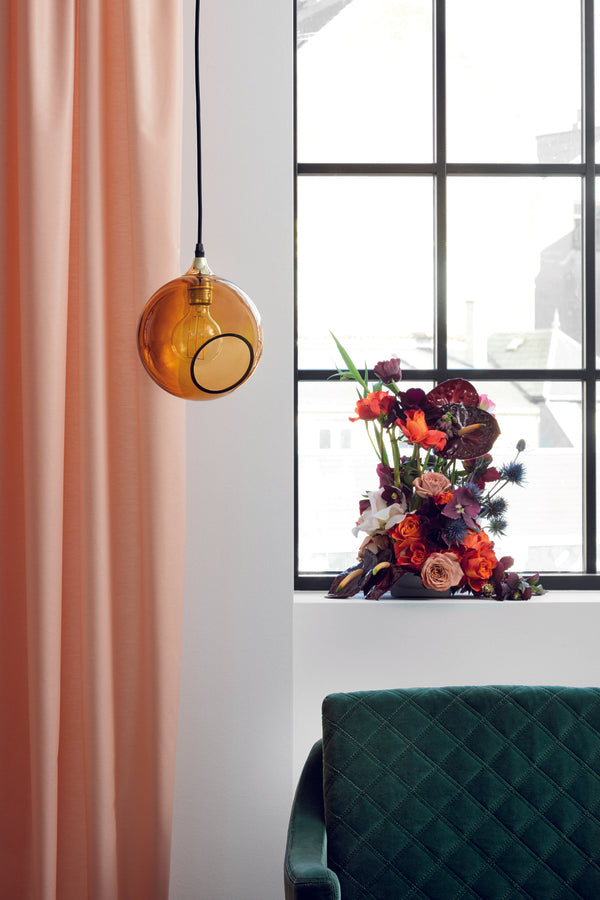 Rund lampa i orange/gult glas med en avskuren bit som ger en skymt av glödlampan. Rosettens sockel är guldfärgad och kabeln är gjord av svart textil.