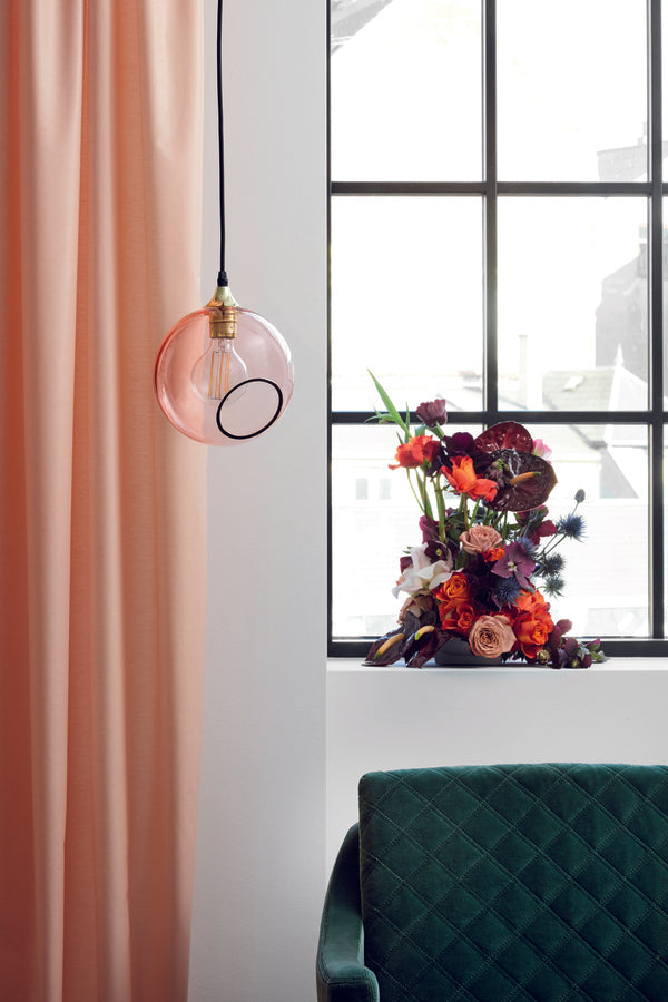 Rund lampa i rosafärgat glas med en avskuren bit med guldkant. Sockeln är guldfärgad och kabeln av svart textil.