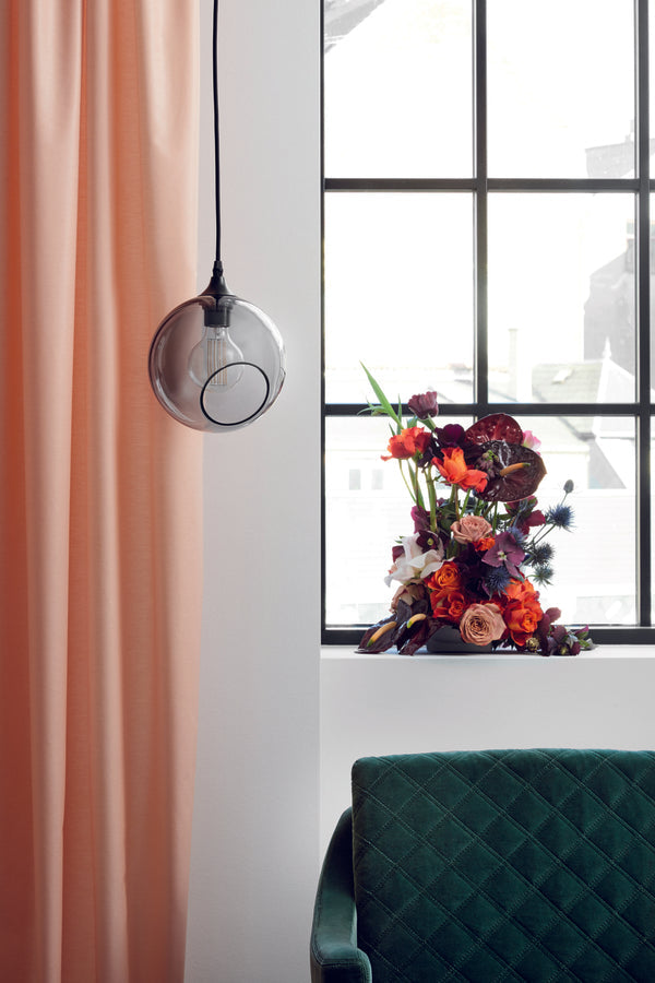 Rund pendellampa i rökfärgat glas med en avskuren bit som ger en skymt av glödlampan. Sockeln är svart och kabeln av svart textil.