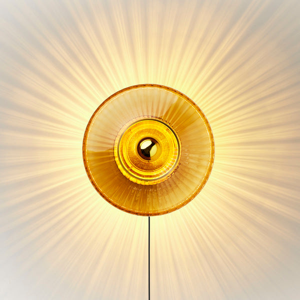 Rund vägglampa i amber (orange/gult) veckat glas som skapar ett ljusspel på väggen.