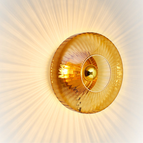 Rund vägglampa i amber (orange/gult) veckat glas som skapar ett ljusspel på väggen.