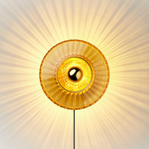 Rund vägglampa i amber (orange/gult) veckat glas som skapar ett ljusspel på väggen.