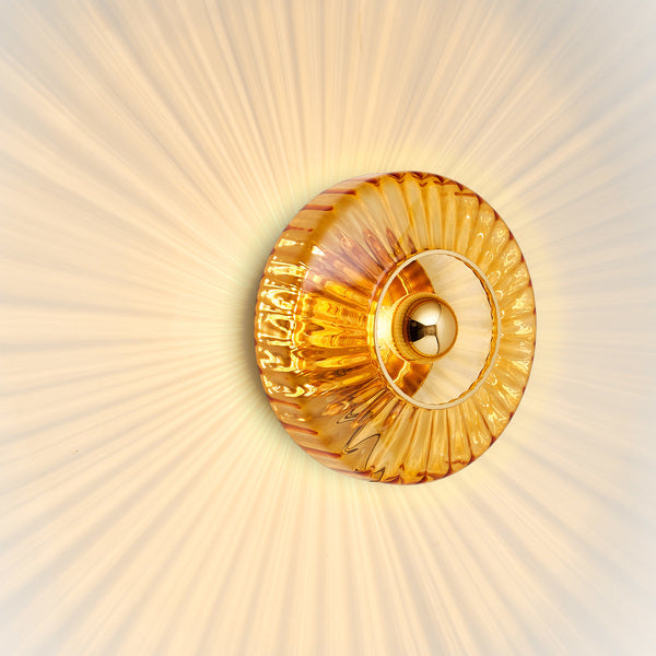 Rund vägglampa i amber (orange/gult) veckat glas som skapar ett ljusspel på väggen.