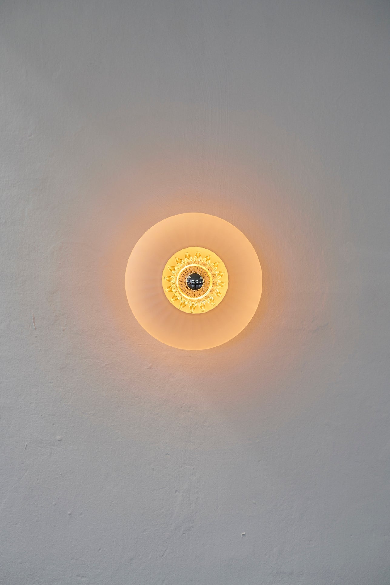 Rund vägglampa i opalfärgat veckat glas som skapar ett ljusspel på väggen.