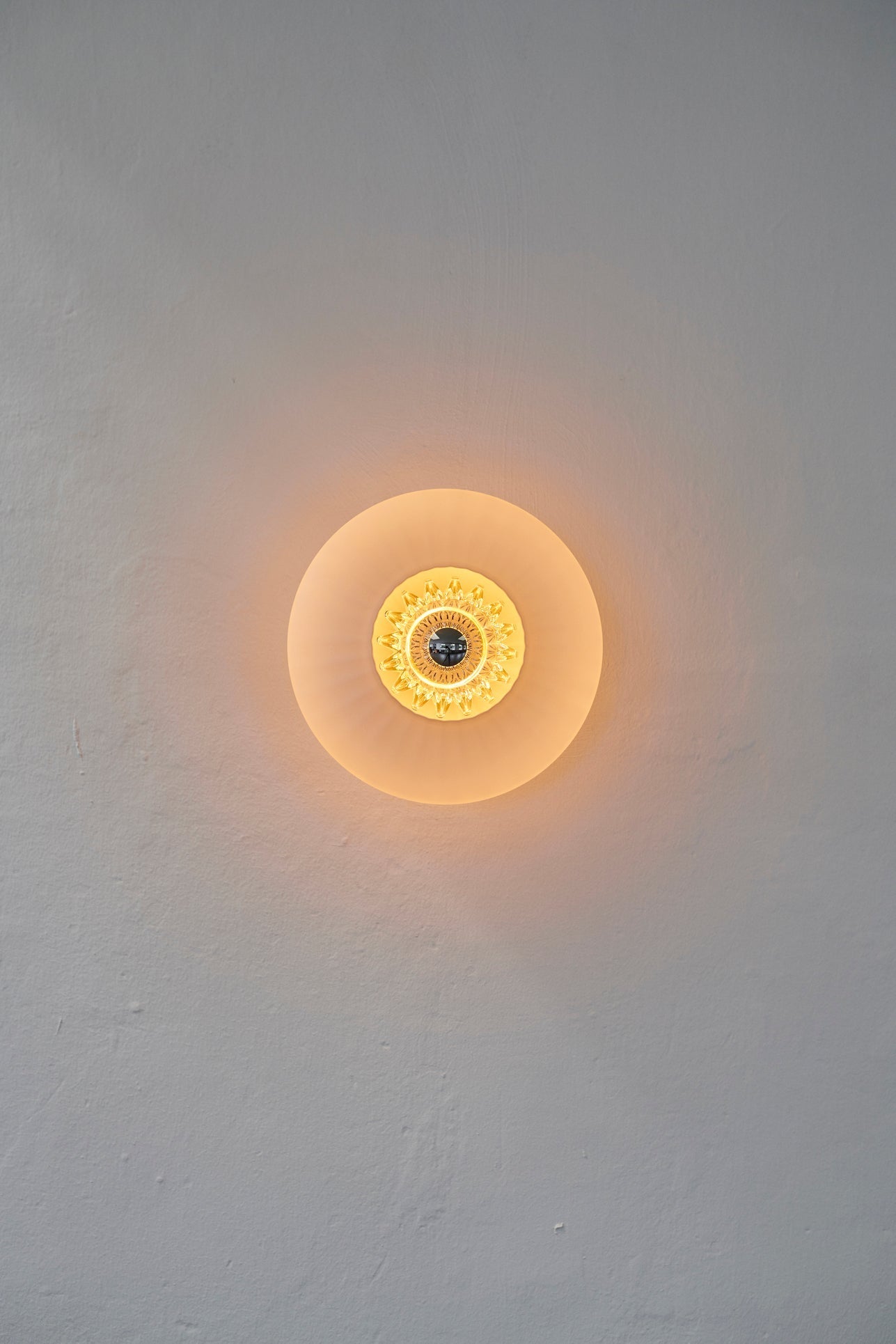 Rund vägglampa i veckat glas i färgen amber som skapar ett ljusspel på väggen.