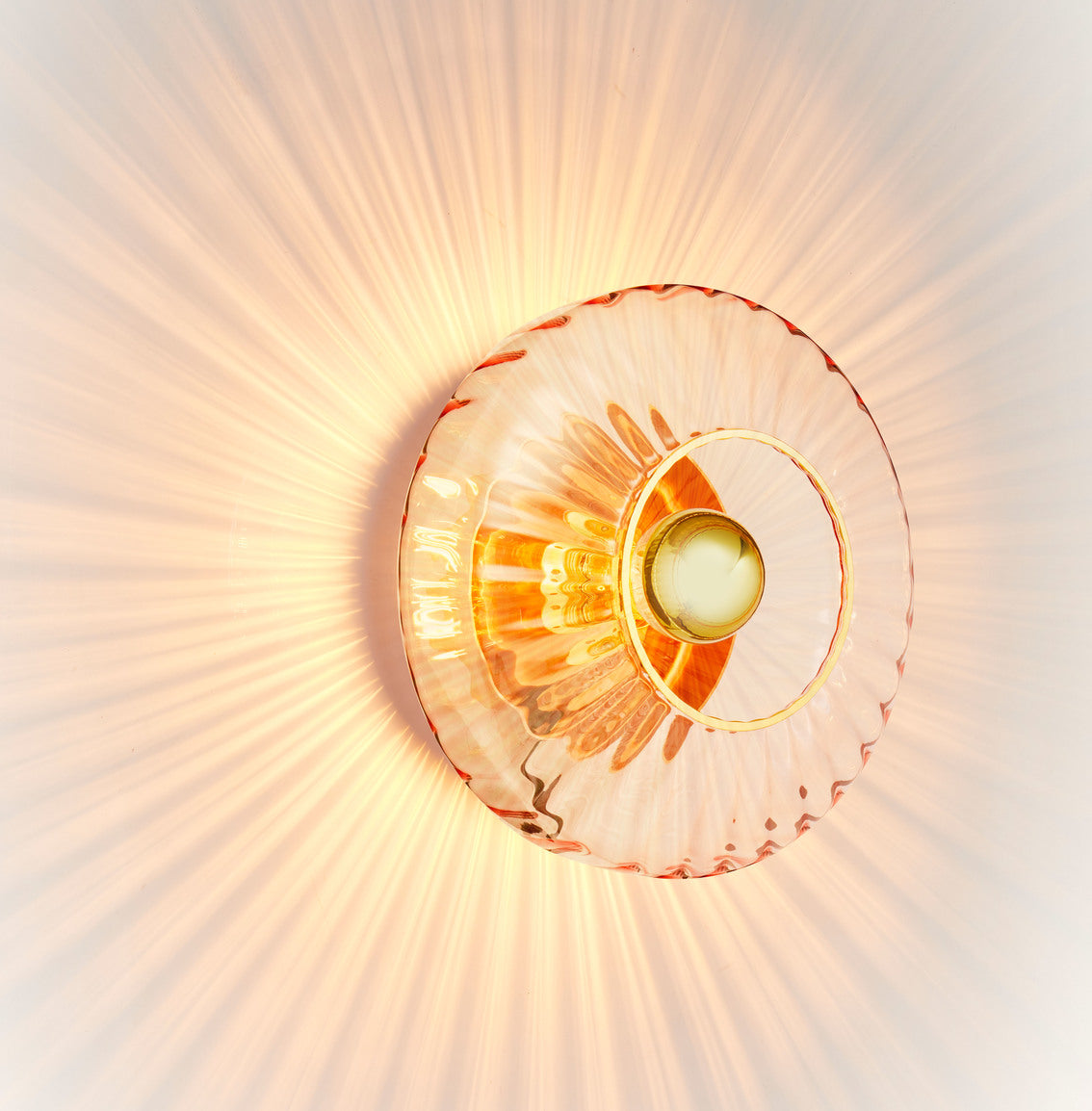 Rund vägglampa i rosa veckat glas som skapar ett ljusspel på väggen.