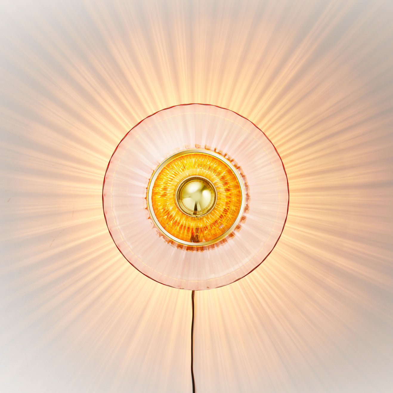 Rund vägglampa i rosa veckat glas som skapar ett ljusspel på väggen.