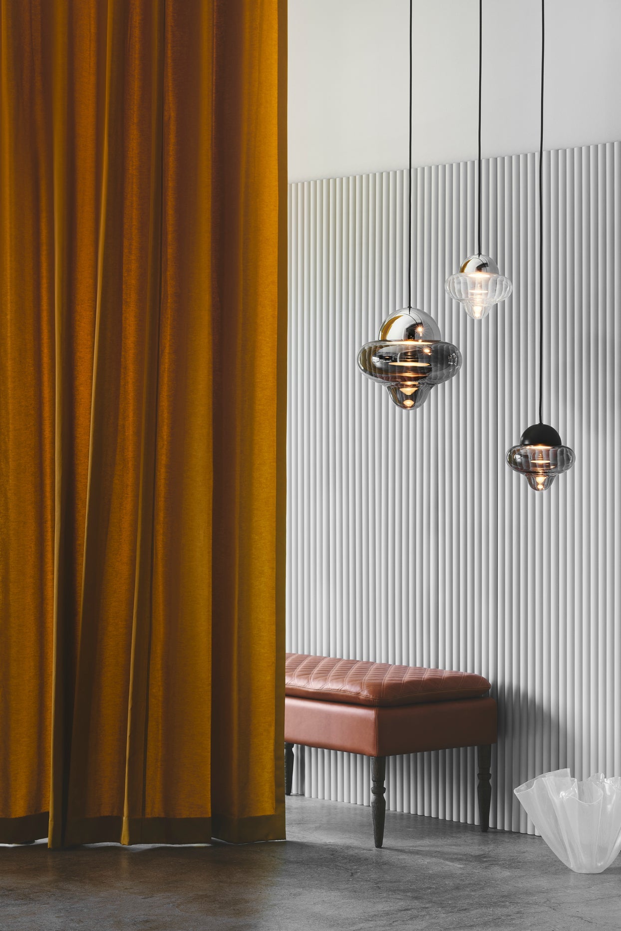 En stor och två små Nutty Clear pendellampor som hänger ner från taket i ett rum med en ljusgrå böljande vägg.