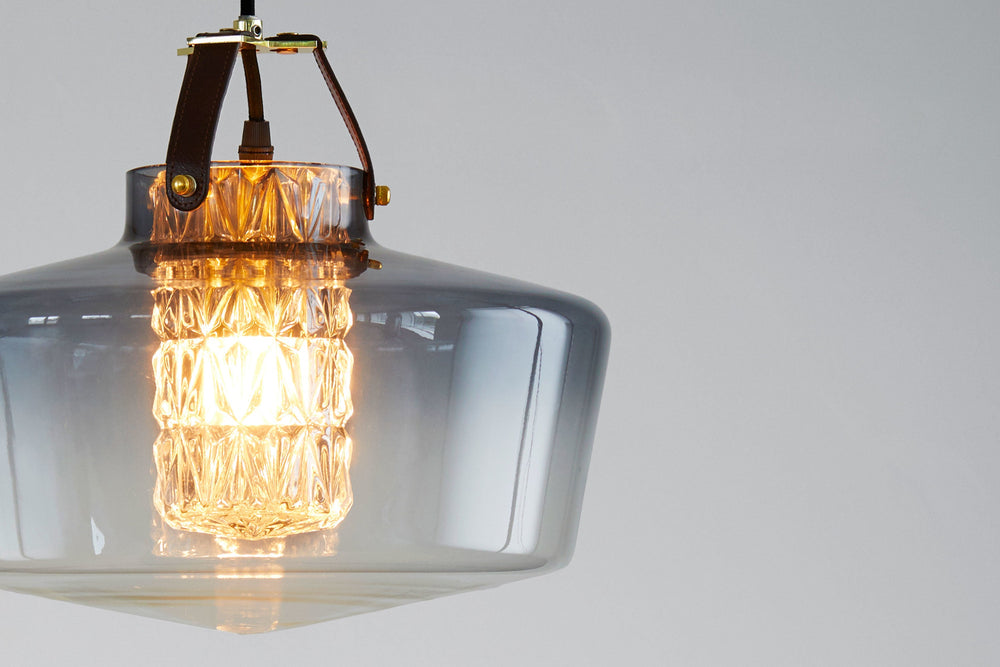 Lampan har en rökfärgad glasskärm i munblåst glas, ett fasettslipat cylinderrör och unika detaljer, bland annat i läder.