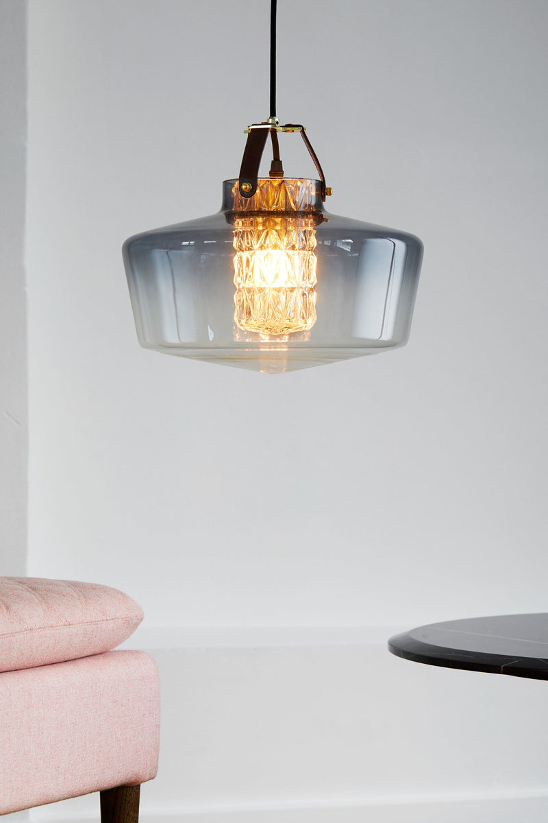 Lampan har en rökfärgad glasskärm i munblåst glas, ett fasettslipat cylinderrör och unika detaljer, bland annat i läder.