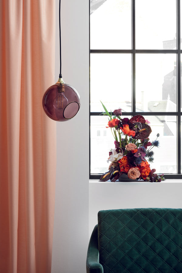 Rund lampa i lila glas med en avskuren bit med guldkant som ger en skymt av glödlampan. Sockel och rosett är guldfärgade och sladden är av svart tyg.