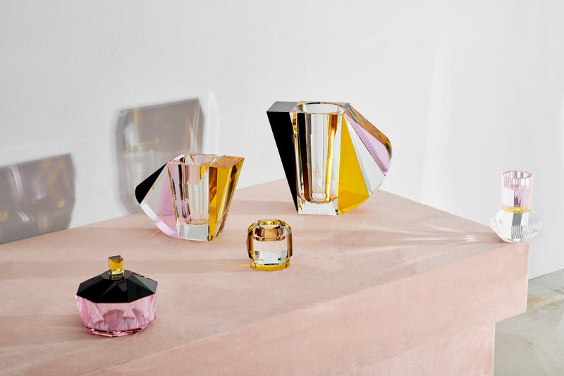 Arrangemang med vaser, ljusstakar och bonbonjär i färgad kristall, på ett rosa bord mot vit bakgrund.