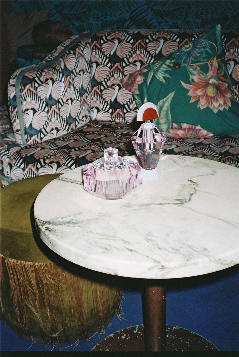 Arrangemang med doftljus i färgad kristallbehållare samt flaska i färgad kristall, stående på ett runt marmorbord med loungemöbler i bakgrunden.