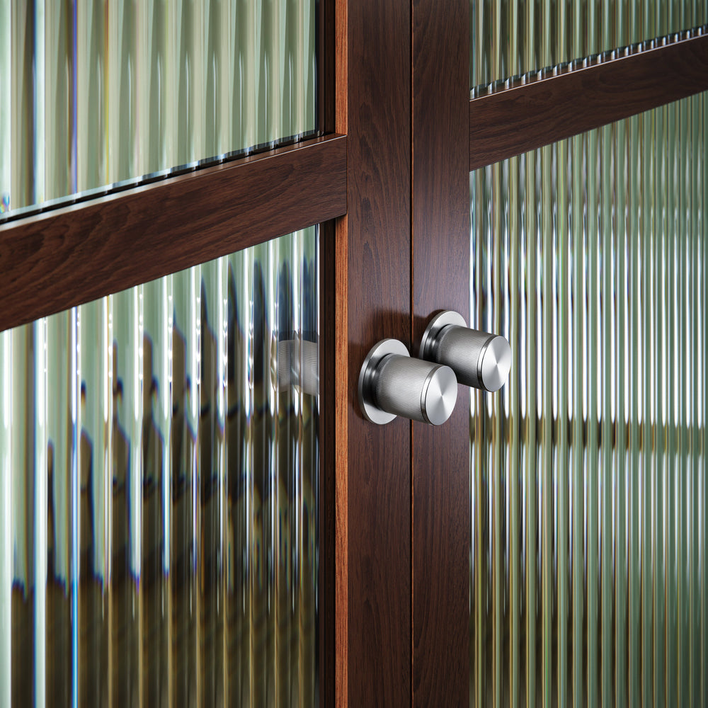 Exklusiv dörrknopp i rostfritt stål med linjerat mönster. Här visas två dörrknoppar monterade på en trädörr med glasruta.