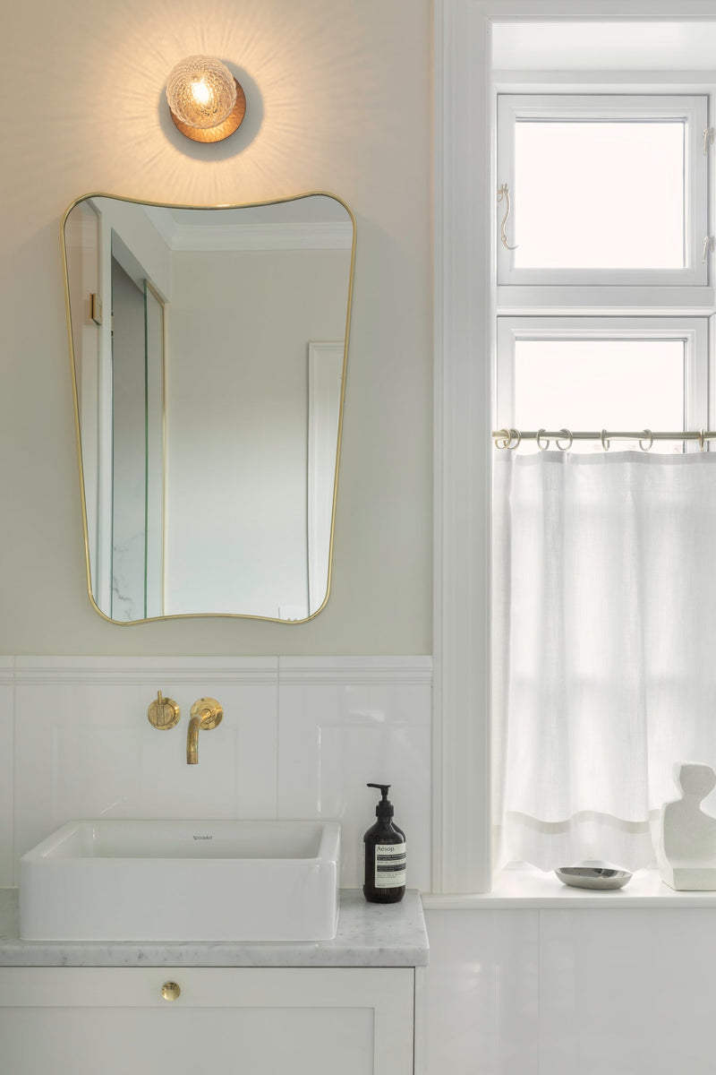 Væglampe med lampeskærm af klart optikglas og gylden fatning, hængende over spejl på badeværelse
