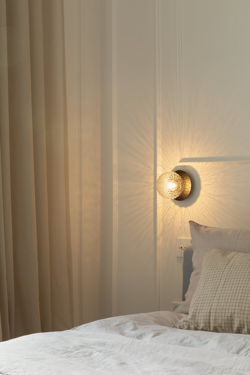 Væglampe med lampeskærm af klart optikglas og gylden fatning, hængende i soveværelse