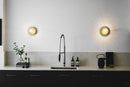 To væglamper med lampeskærme af klart optikglas og gylden fatning, over køkkenbord