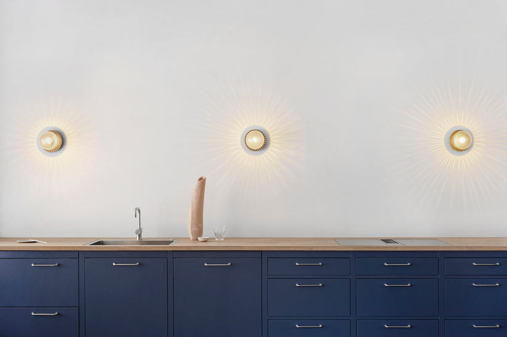 Tre væglamper med lampeskærme af klart optikglas og gylden fatning, over blåt køkken