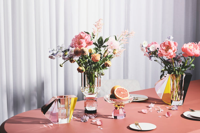 Arrangemang med vaser och en skål i färgad kristall, på ett korallfärgat bord med en vit gardin i bakgrunden