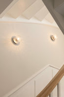 To væglampe med lampeskærme af klart optikglas og sølvfarvet fatning, hængende på væg langs trappe.