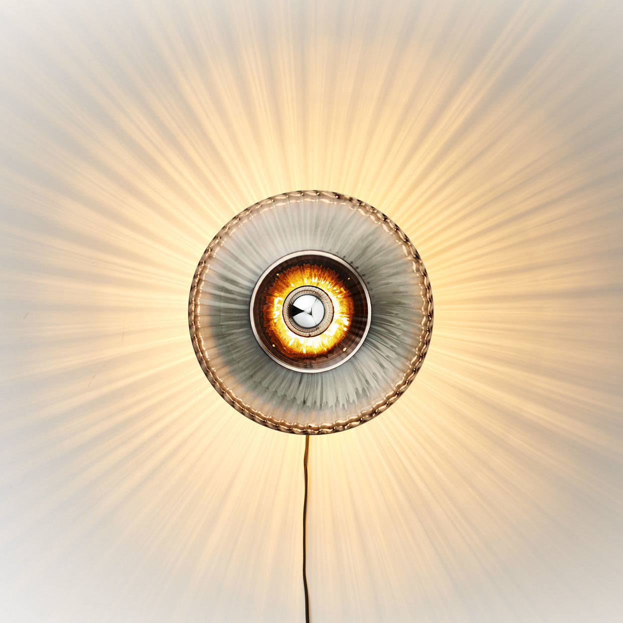 Rund vägglampa i rökfärgat veckat glas som skapar ett ljusspel på väggen. 