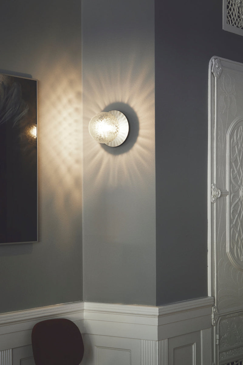 Væglampe med lampeskærm af klart optikglas og sølvfarvet fatning, hængende i dagligstuemiljø