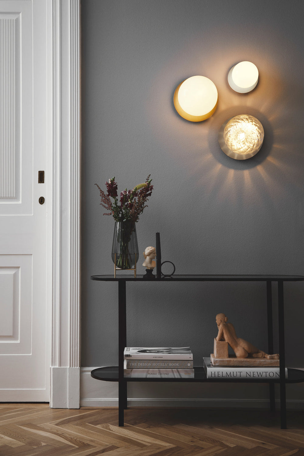 Tre væglamper med lampeskærme af klart optikglas og sølvfarvet fatning, hængende på væg i dagligstuemiljø
