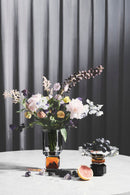 Arrangemang med vas i klar och bärnstensfärgad kristall med blommor i och skål med frukt, på marmorbord och gardin i bakgrunden.