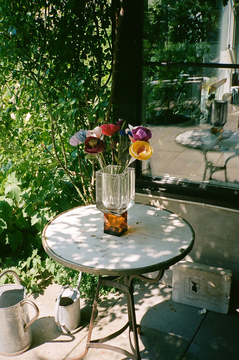 Vas i klar och bärnstensfärgad kristall med blommor i, på ett utebord och med växter i bakgrunden.