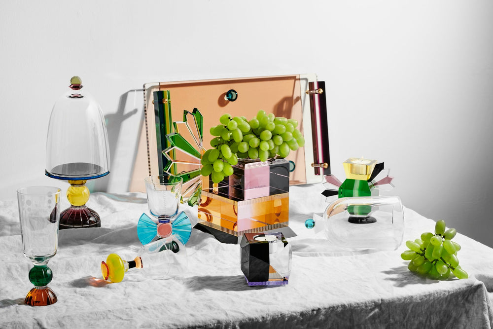 Arrangemang med olika ljusstakar, glas och skålar i färgad kristall, på ett middagsbord med duk och frukt.
