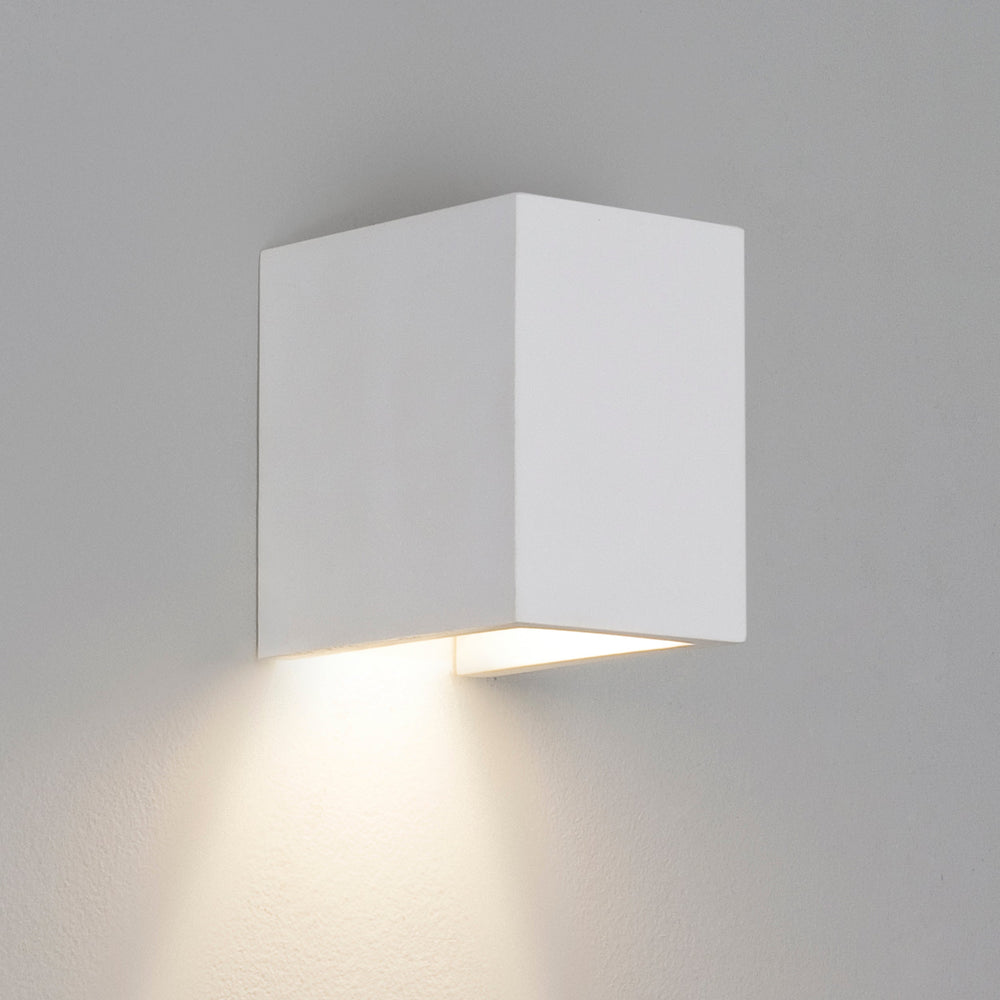En liten vit, enkel vägglampa med kantig form tillverkad av gips. Med nedåtriktat ljus.