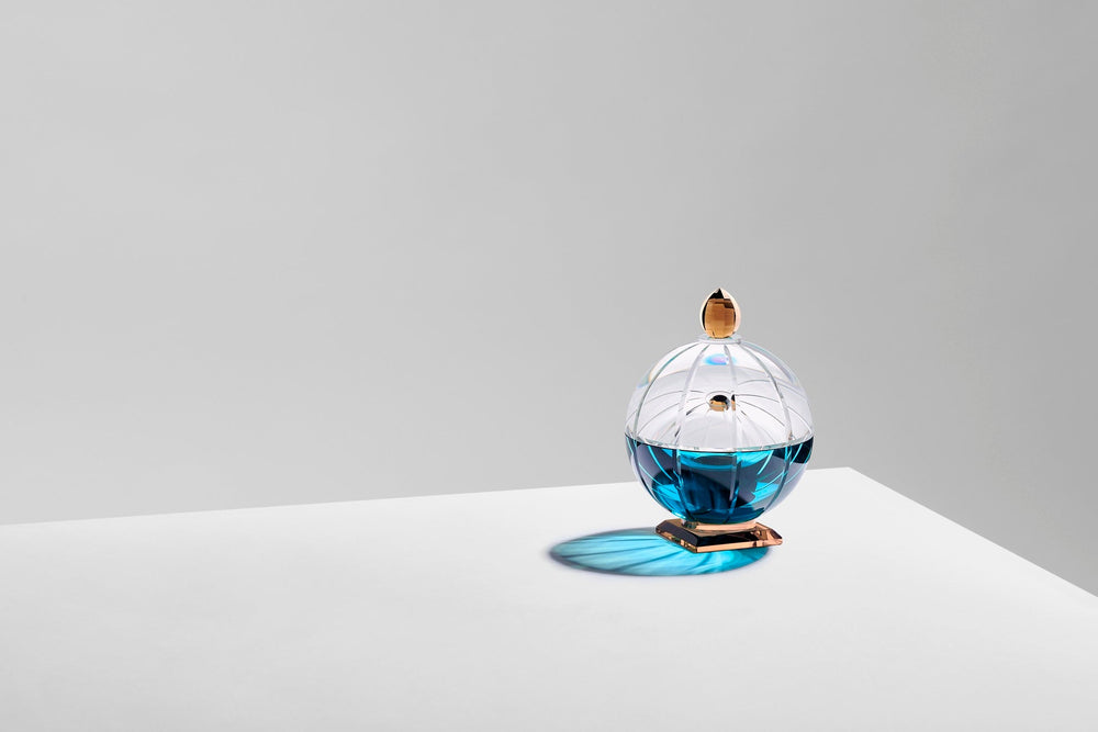 Doftljus i en rund behållare av klar, blå och brun kristall, på ett vitt bord mot grå bakgrund