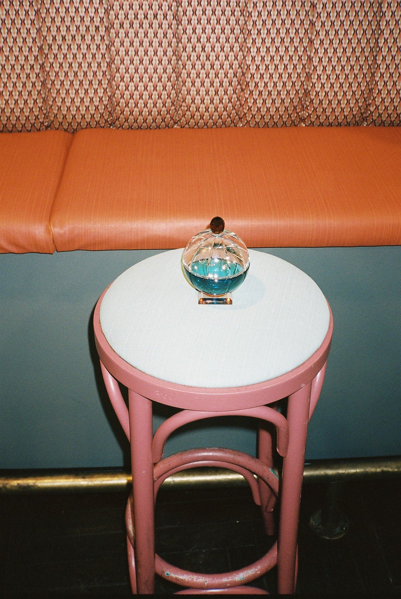 Doftljus i en rund behållare av klar, blå och brun kristall, stående på en taburett med en soffa i bakgrunden
