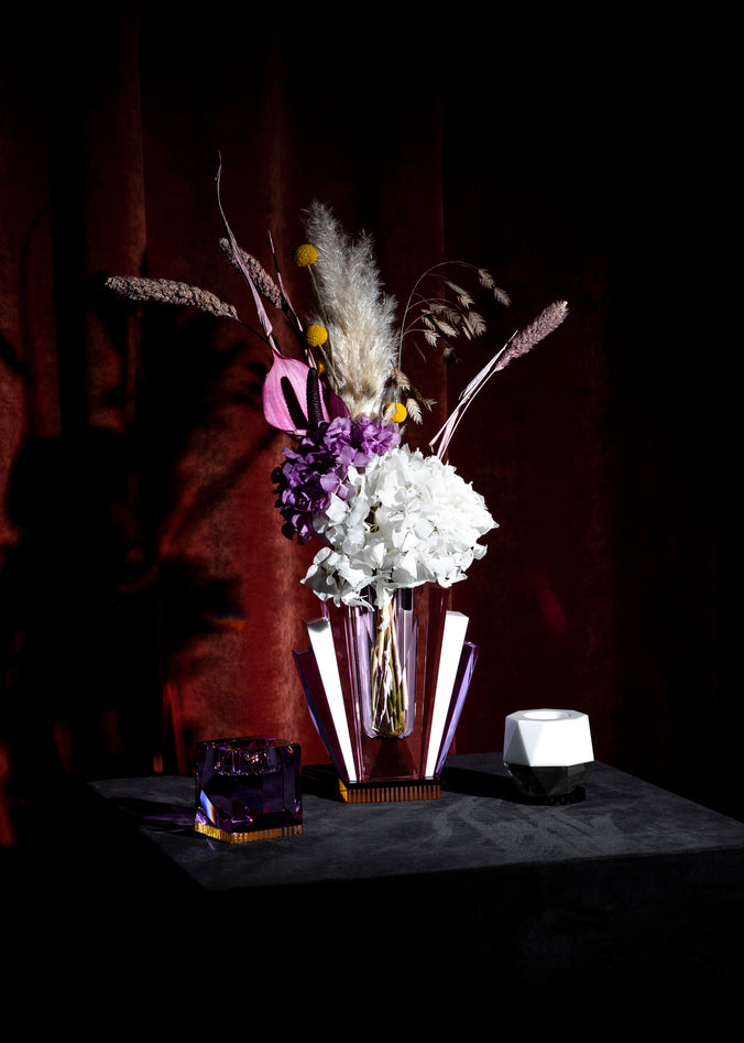 Vas i klar, rosa, lila och gul kristall med blommor i, stående intill två värmeljusstakar i kristall på ett svart bord mot röd bakgrund.