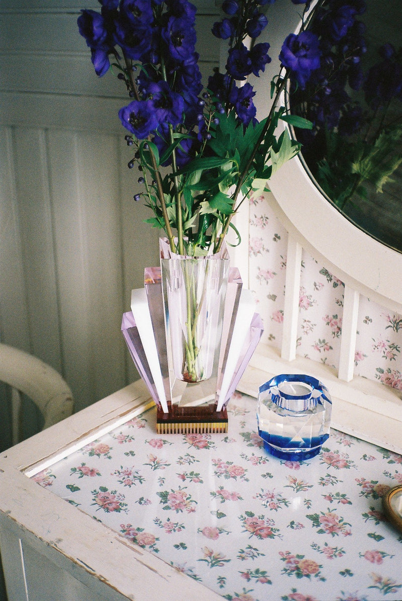 Vas i klar, rosa, lila och gul kristall med blommor tillsammans med värmeljusstake på byrå.