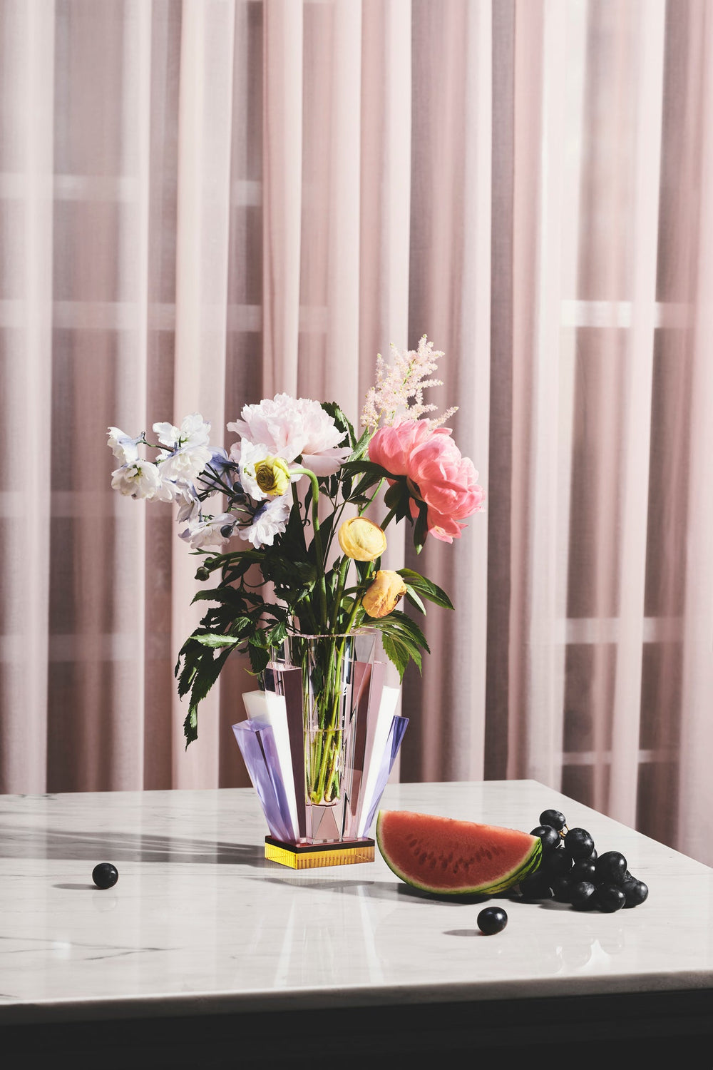 Vas i klar, rosa, lila och gul kristall med blommor i, stående på marmorbord tillsammans med frukt och med en rosa gardin i bakgrunden.