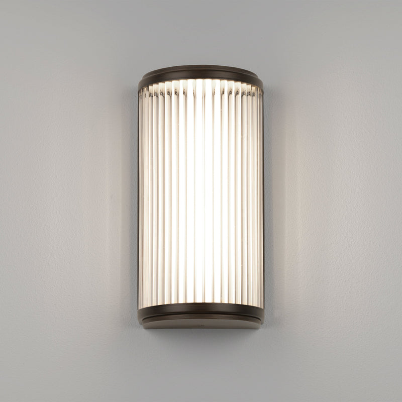 Lampa med topp, botten och baksida i bronsfärgat stål och en glasskärm med nedåtgående linjer.